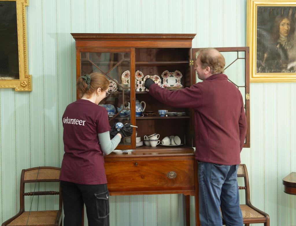 Curators carefully cleaning old and ornate utensils and cups displayed in a cabinet, preserving delicate historical items with attention and care.